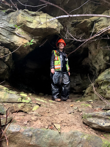 Image of Caleigh Shoot standing outside a cavern in Coos Bay. Caleigh is wearing an orange hardhat and safety vest, and heavy-duty gear. The cavern entrance has a dirt path leading up to it, and features a rocky entrance.