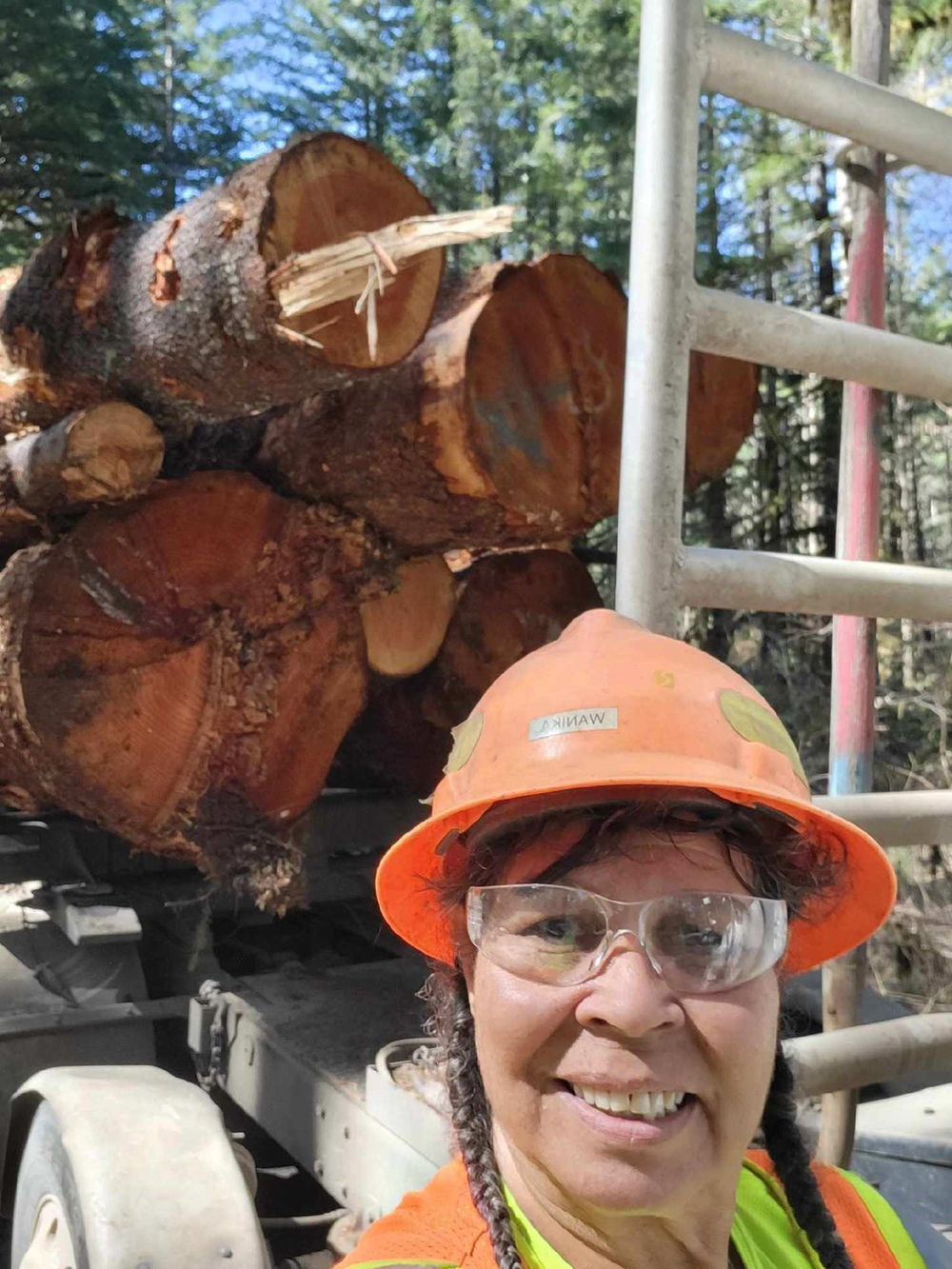 Image of Wanika Doggett with a load of logs on her truck. Wanika is wearing an orange safety help and glasses, as well as a bright safety vest. Behind her, on the bed of her truck, are a number of cut logs ready for transport.
