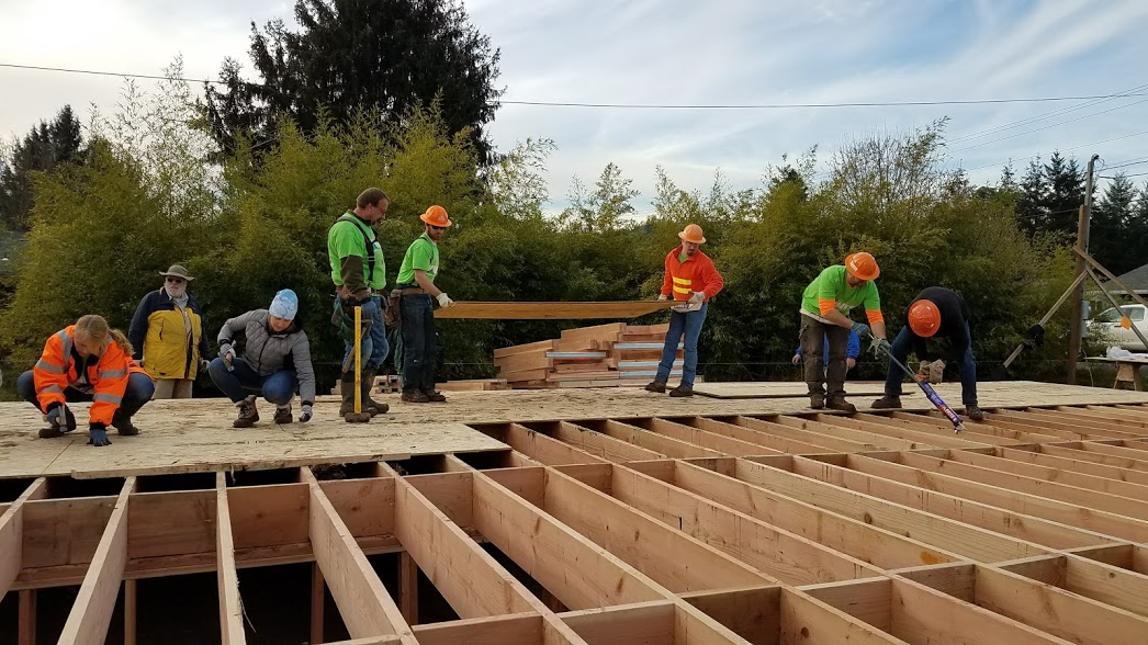 An image of construction on a Habitat for Humanity site, with workers on a work site moving a panel of plywood and others hammer nails.